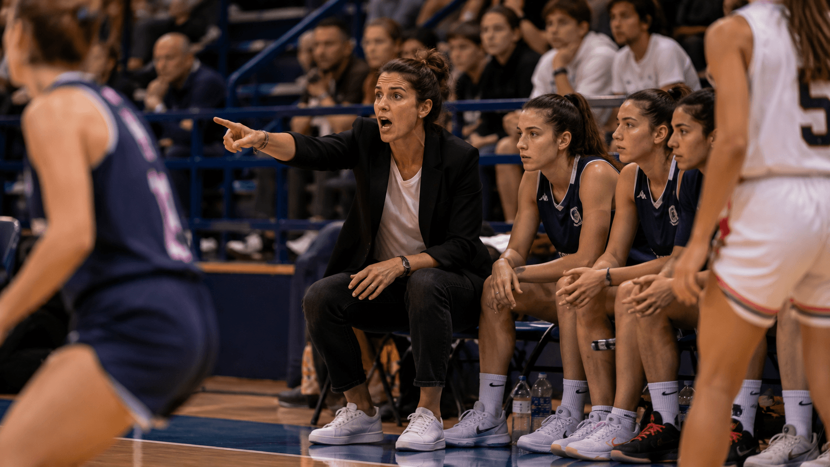 Une coach appelle un système depuis le banc, le bras tendu vers le terrain, pendant que ses remplaçantes suivent l'action.